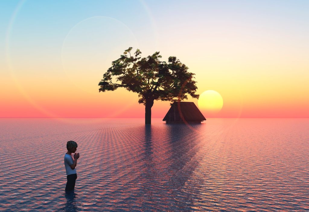 A boy standing in water to his knees that. The water is everywhere. There is a tree and and a house partially submerged in the the distance.