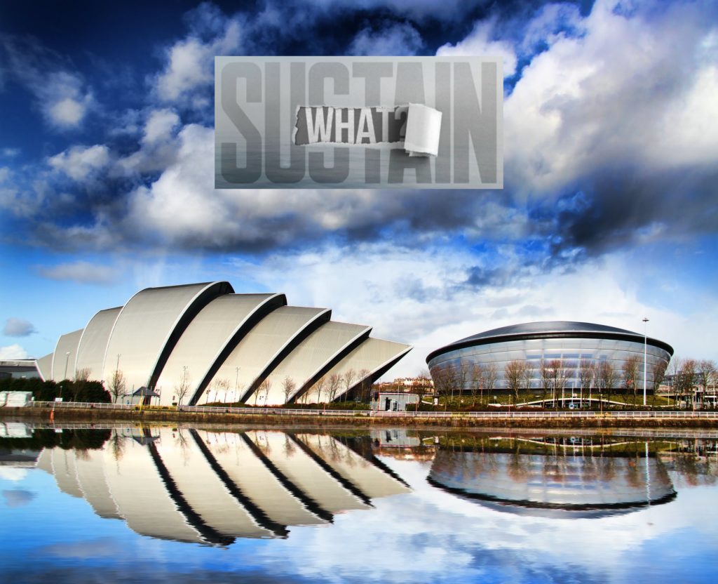 The skyline of the COP26 venue in Glasgow, Scotland. It is reflected in a lake in the foreground. The Sustain What logo is superimposed.