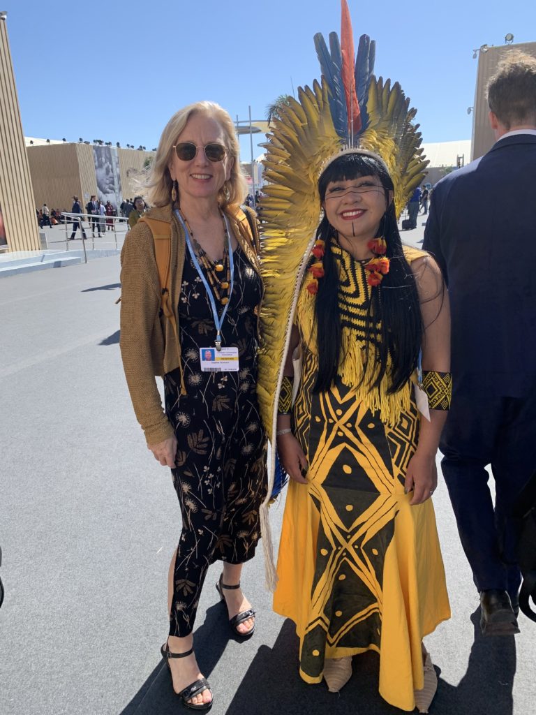 Daphne at COP27 with an indigenous leader. The indigenous woman is stressed in colorful yellow and black patterns with a headdress of yellow blue and red feathers.